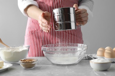 Photo of Making cinnamon rolls. Woman sieving flour at gray marble table, closeup