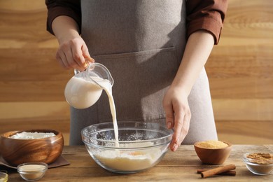 Photo of Making cinnamon rolls. Woman pouring milk into bowl at wooden table, closeup