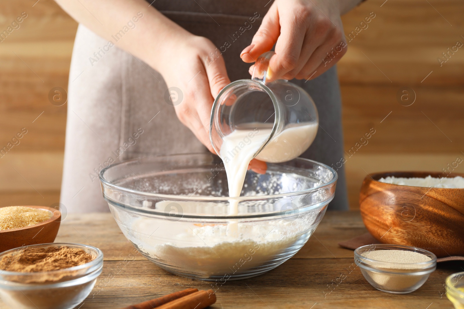 Photo of Making cinnamon rolls. Woman pouring milk into bowl at wooden table, closeup