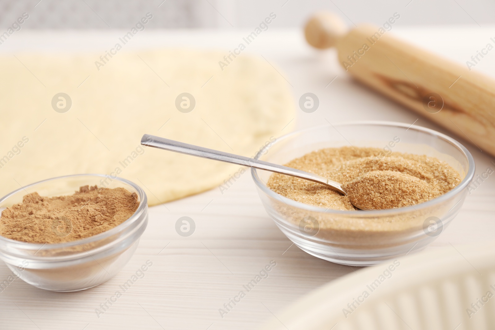 Photo of Making cinnamon rolls. Spice, sugar, spoon, dough and rolling pin on white wooden table, closeup