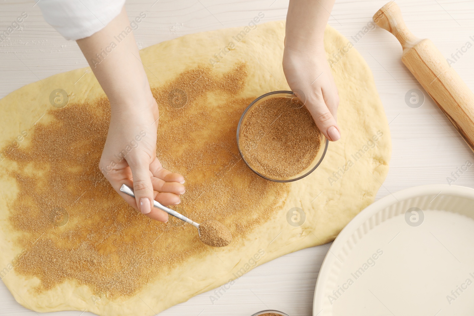 Photo of Making cinnamon rolls. Woman adding spice, with sugar into dough at white wooden table, top view