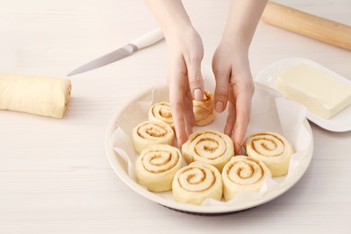 Photo of Woman putting cinnamon roll into baking dish at white wooden table, closeup