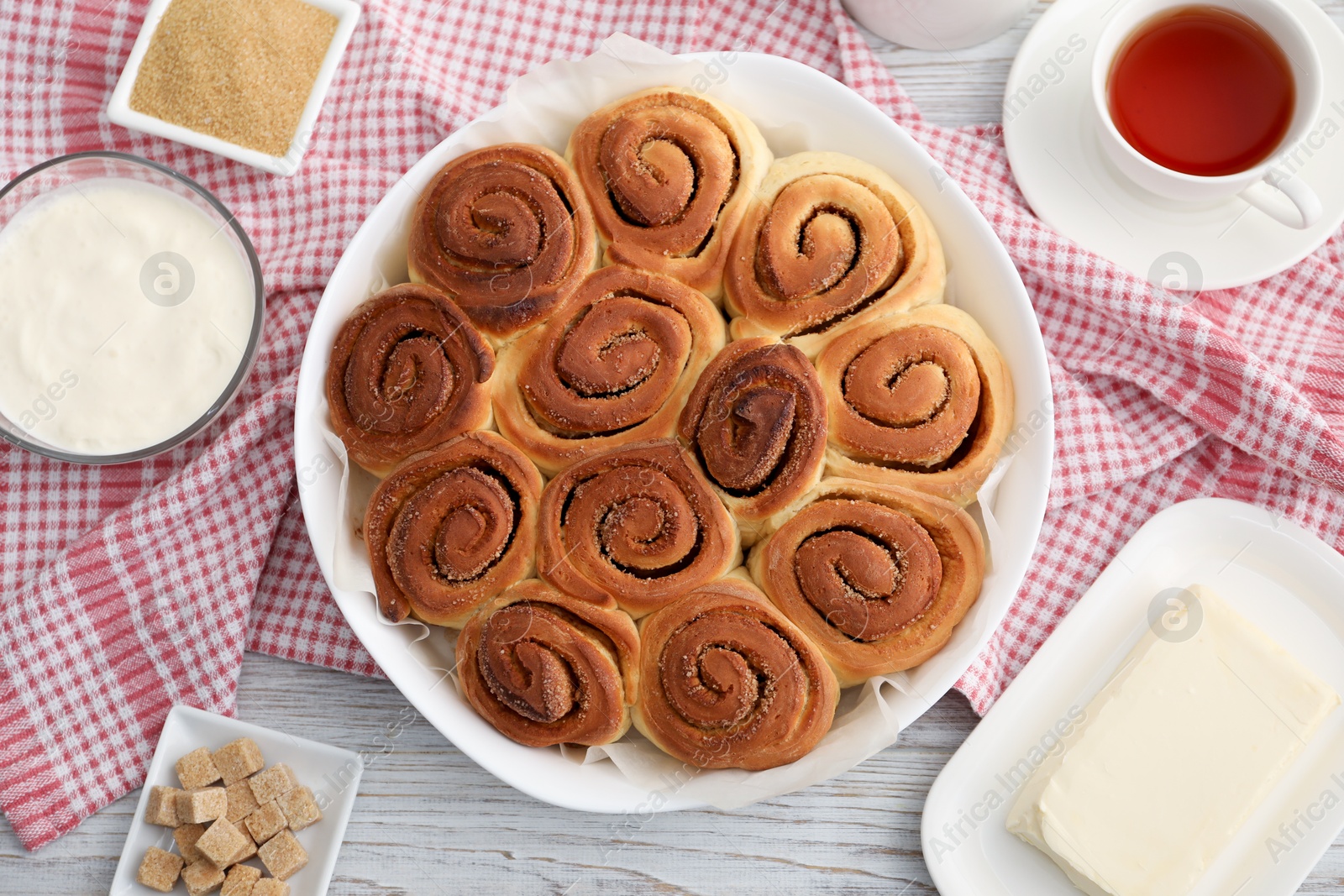 Photo of Freshly baked cinnamon rolls, tea and ingredients on light wooden table, flat lay