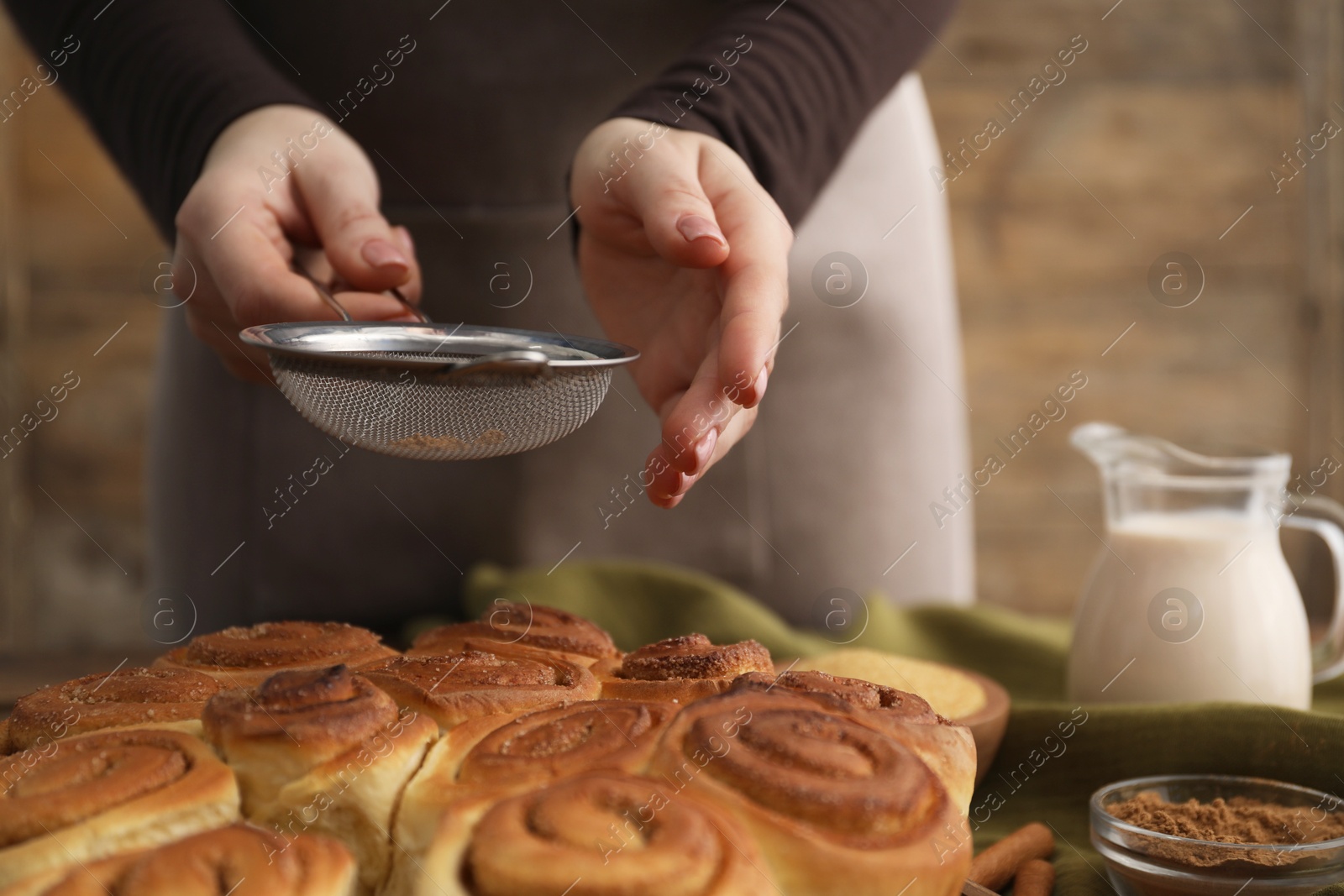 Photo of Woman with sieve adding cinnamon powder onto freshly baked rolls at table, closeup