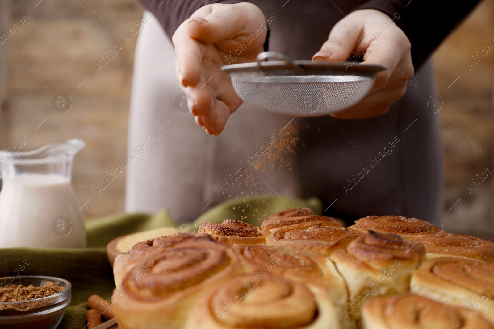 Photo of Woman with sieve adding cinnamon powder onto freshly baked rolls at table, closeup