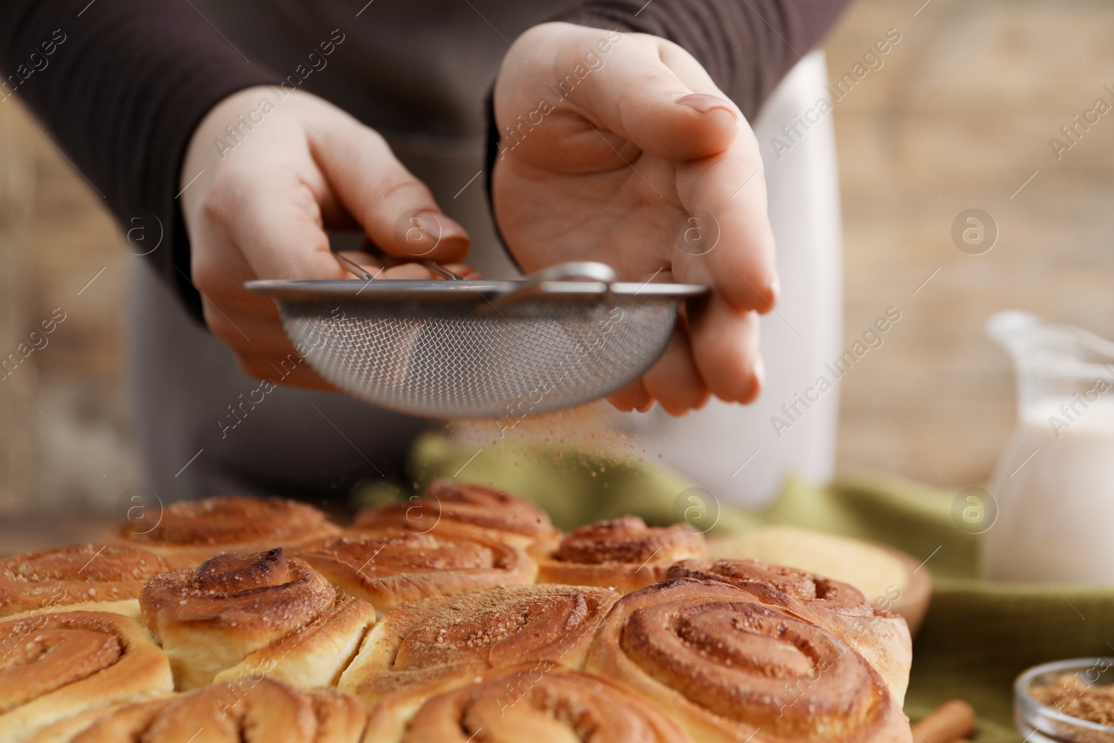 Photo of Woman with sieve adding cinnamon powder onto freshly baked rolls at table, closeup