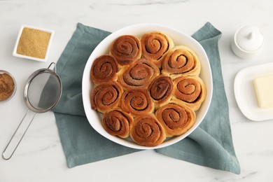 Photo of Tasty cinnamon rolls, ingredients and sieve on white marble table, flat lay