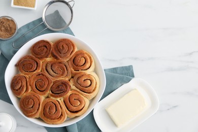 Photo of Tasty cinnamon rolls, ingredients and sieve on white marble table, flat lay. Space for text