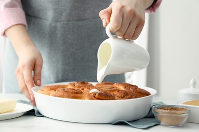 Photo of Woman adding frosting onto freshly baked cinnamon rolls at white table indoors, closeup