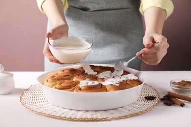 Photo of Woman spreading frosting onto freshly baked cinnamon rolls at white table against pink background, closeup