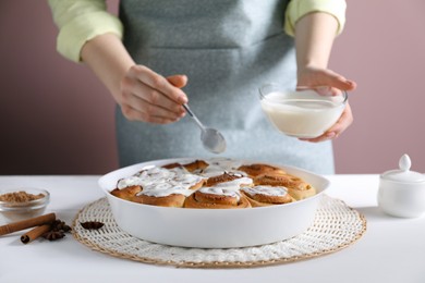 Photo of Woman spreading frosting onto freshly baked cinnamon rolls at white table against pink background, closeup