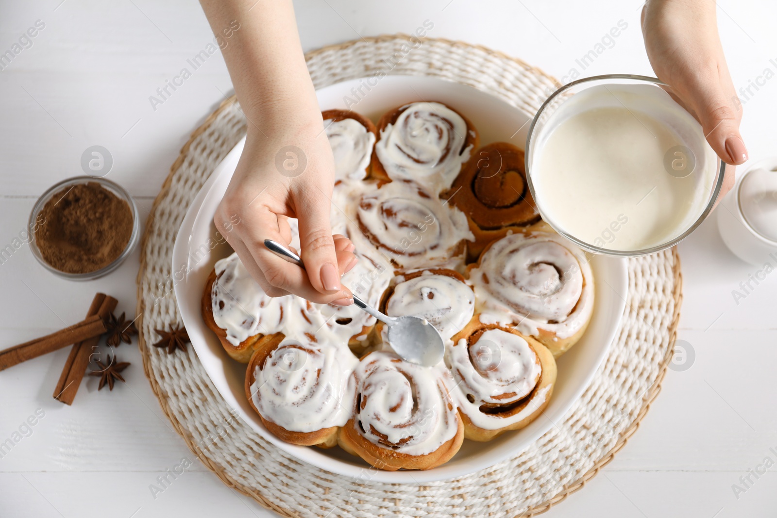 Photo of Woman spreading frosting onto freshly baked cinnamon rolls at white wooden table, top view