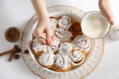 Photo of Woman spreading frosting onto freshly baked cinnamon rolls at white wooden table, top view