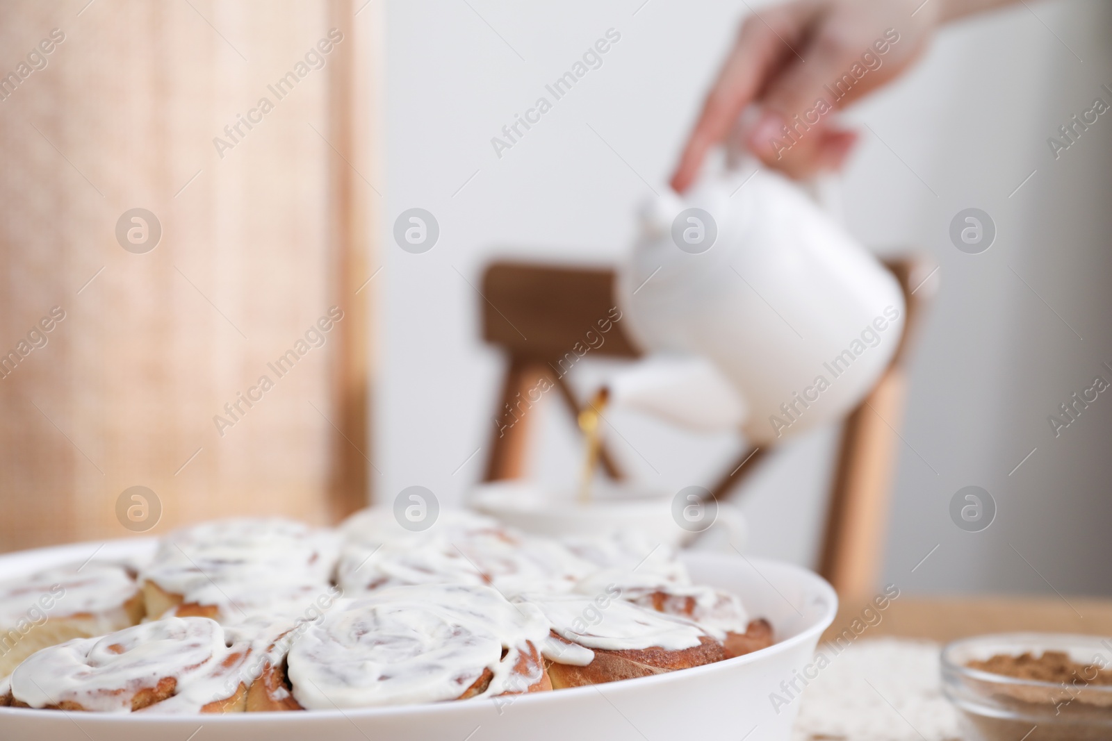 Photo of Delicious frosted cinnamon rolls and woman with teapot at table indoors, selective focus. Space for text