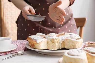 Photo of Woman with sieve adding cinnamon powder onto freshly baked rolls at table indoors, closeup