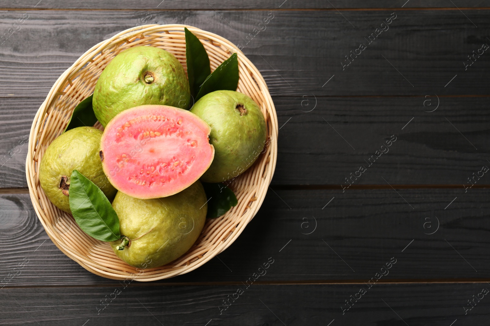 Photo of Fresh whole and cut guava fruits in wicker basket on black wooden table, top view. Space for text