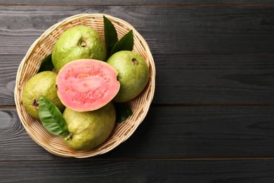 Fresh whole and cut guava fruits in wicker basket on black wooden table, top view. Space for text