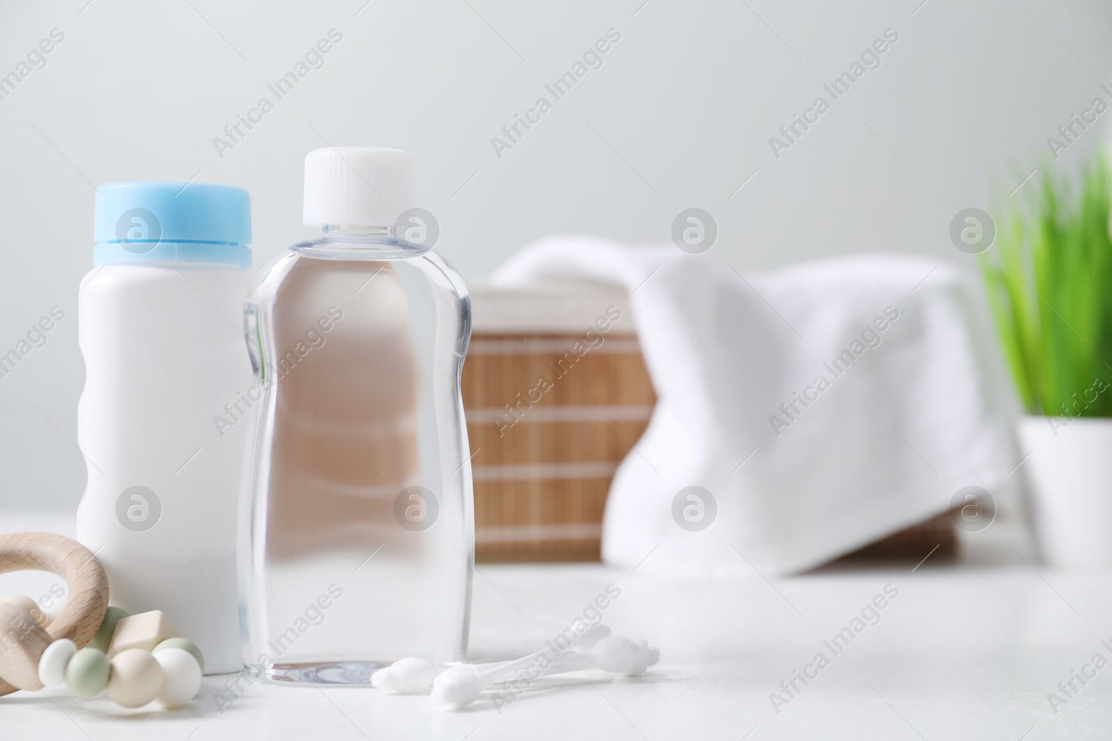 Baby oil, powder, cotton swabs and teether on white table against blurred background. Space for text Photo of Baby oil, powder, cotton swabs and teether on white table against blurred background. Space for text