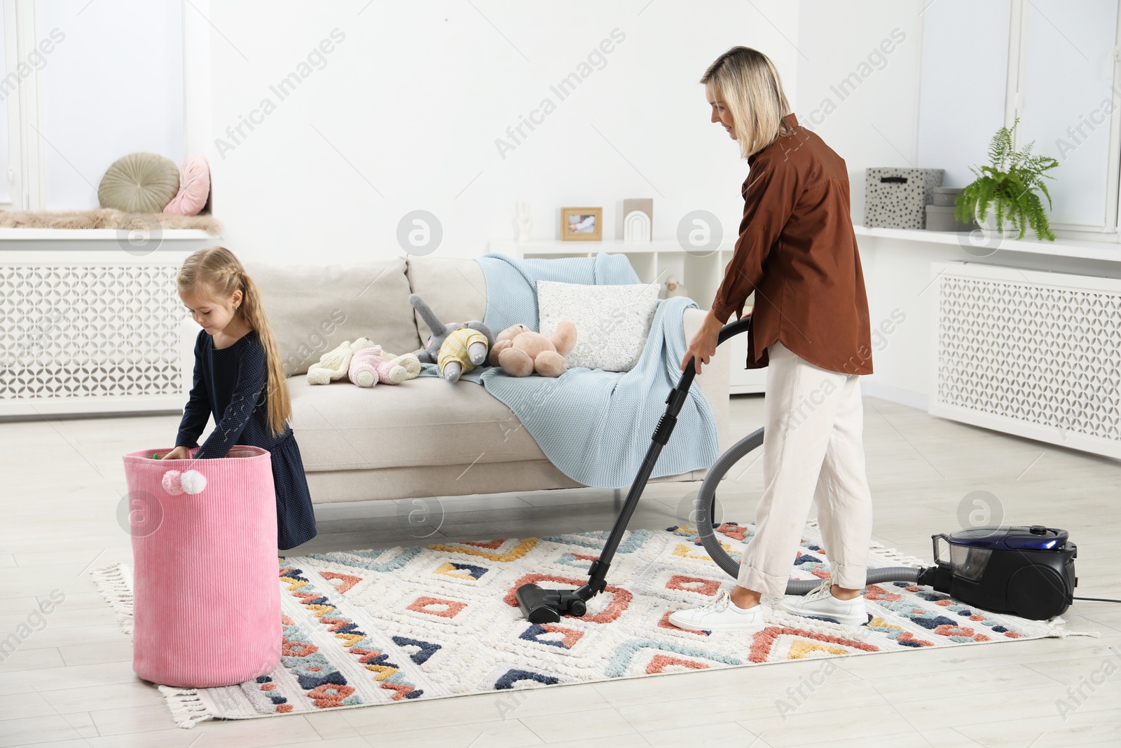 Photo of Little helper. Girl putting away toys while her mother vacuuming at home