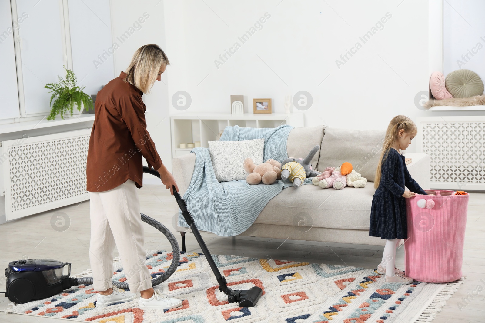 Photo of Little helper. Girl putting away toys while her mother vacuuming at home