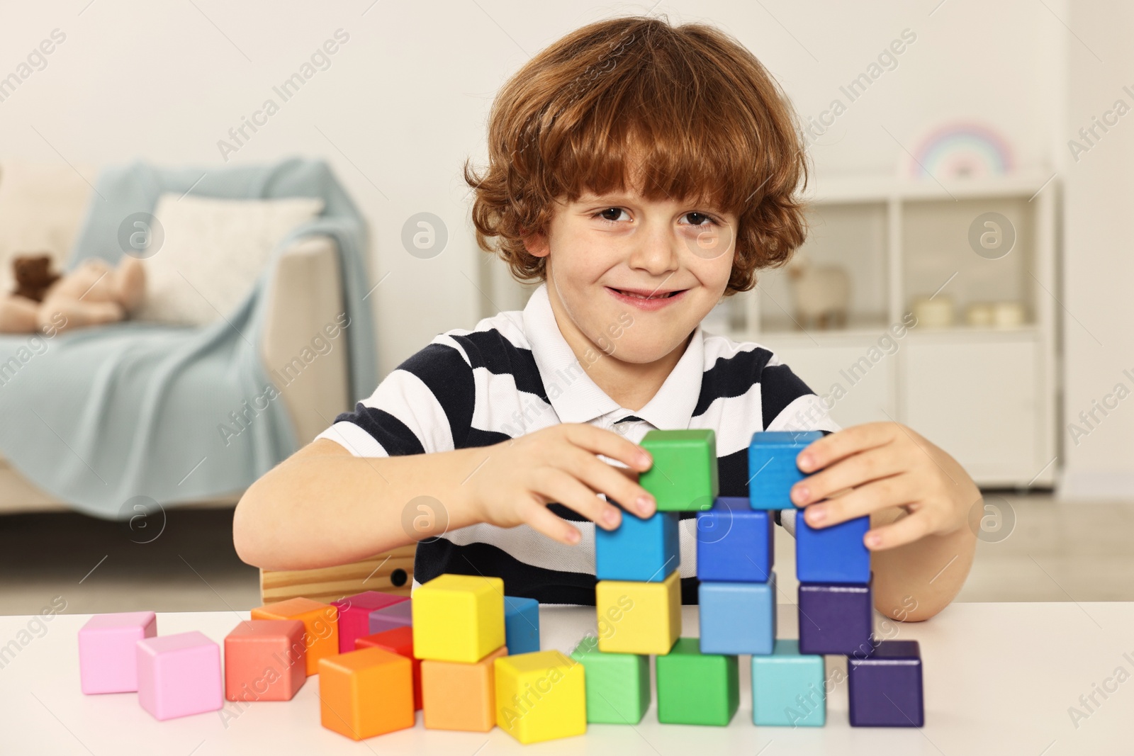 Photo of Little boy stacking colorful cubes at white table indoors