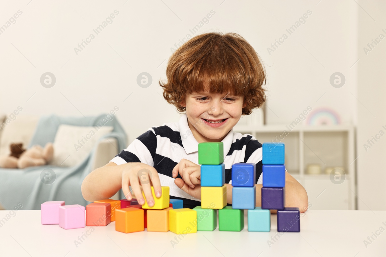 Photo of Little boy stacking colorful cubes at white table indoors