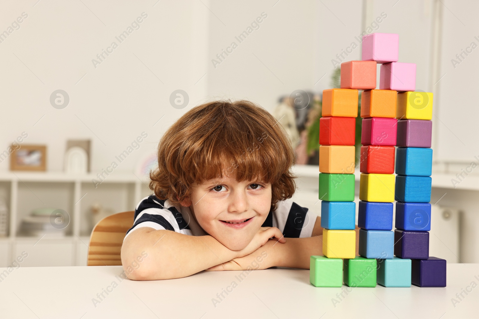 Photo of Little boy playing with stacked colorful cubes at white table indoors