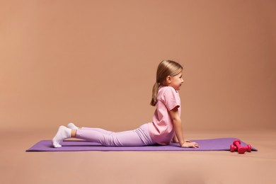 Photo of Cute little girl exercising on fitness mat against dark beige background