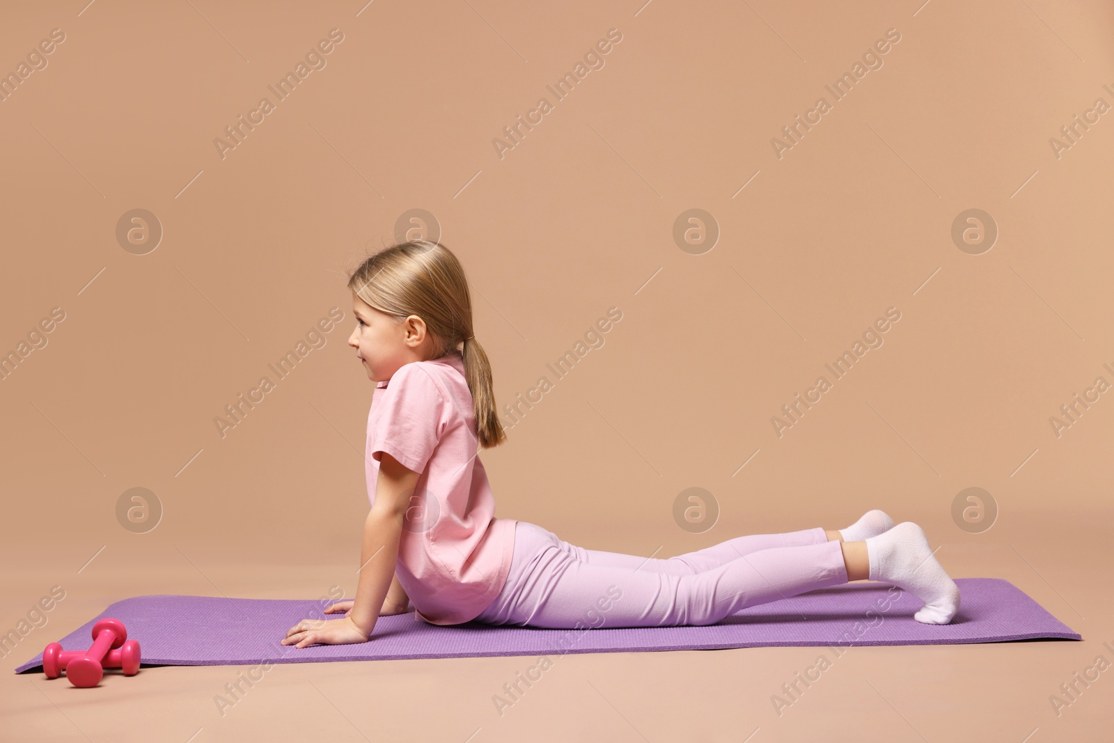 Photo of Cute little girl exercising on fitness mat against dark beige background