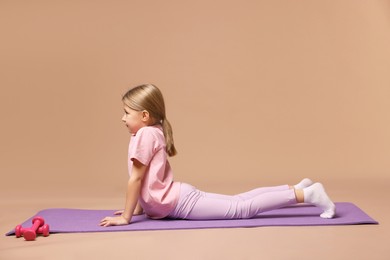 Photo of Cute little girl exercising on fitness mat against dark beige background