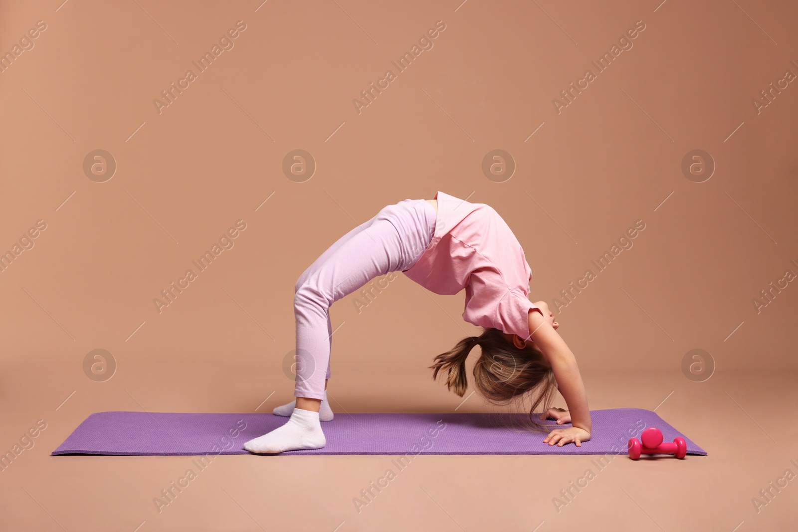 Photo of Cute little girl exercising on fitness mat against dark beige background