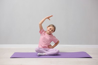 Photo of Cute little girl exercising on fitness mat near grey wall indoors