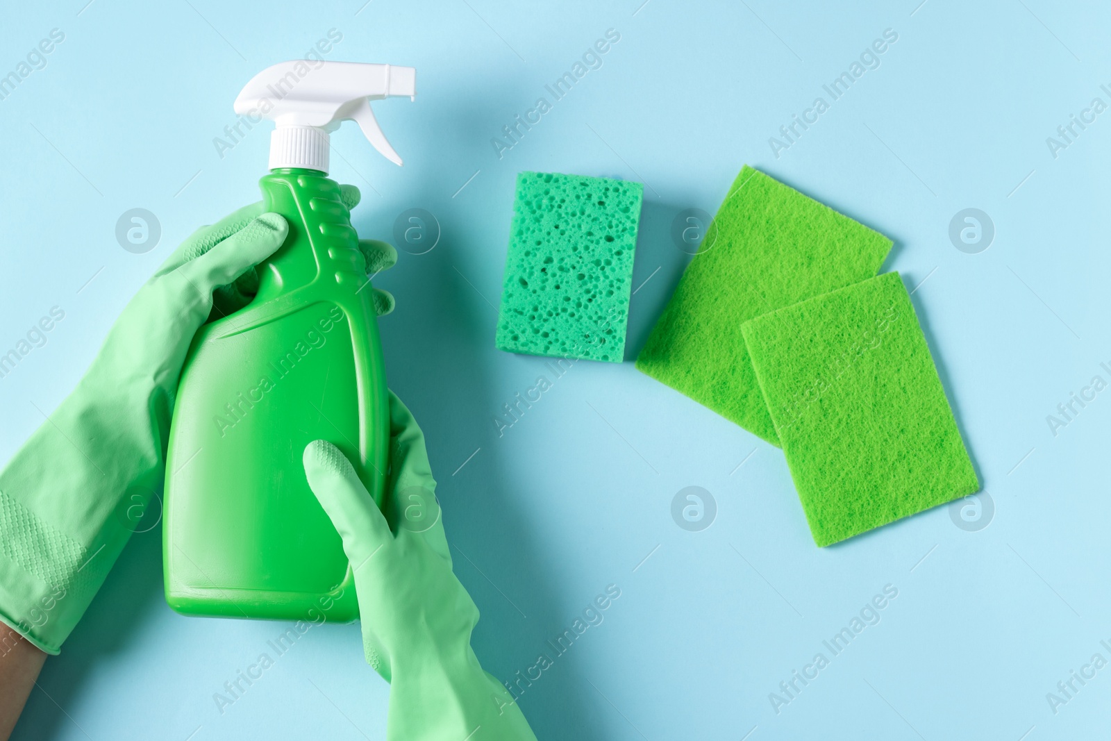 Photo of Woman with bottle of cleaning product and sponges on light blue background, top view