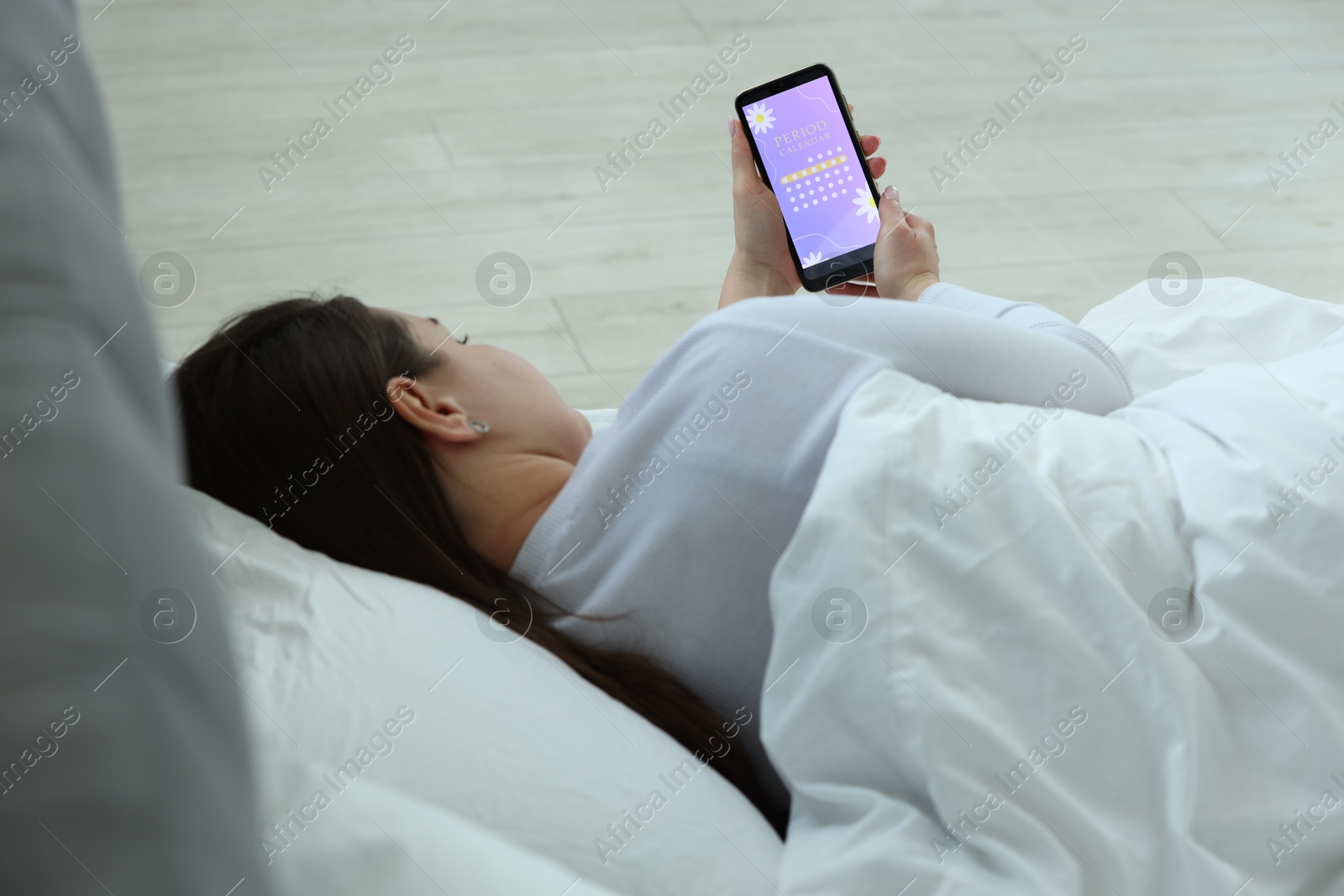 Photo of Menstruation. Woman using period calendar application on smartphone in bed indoors