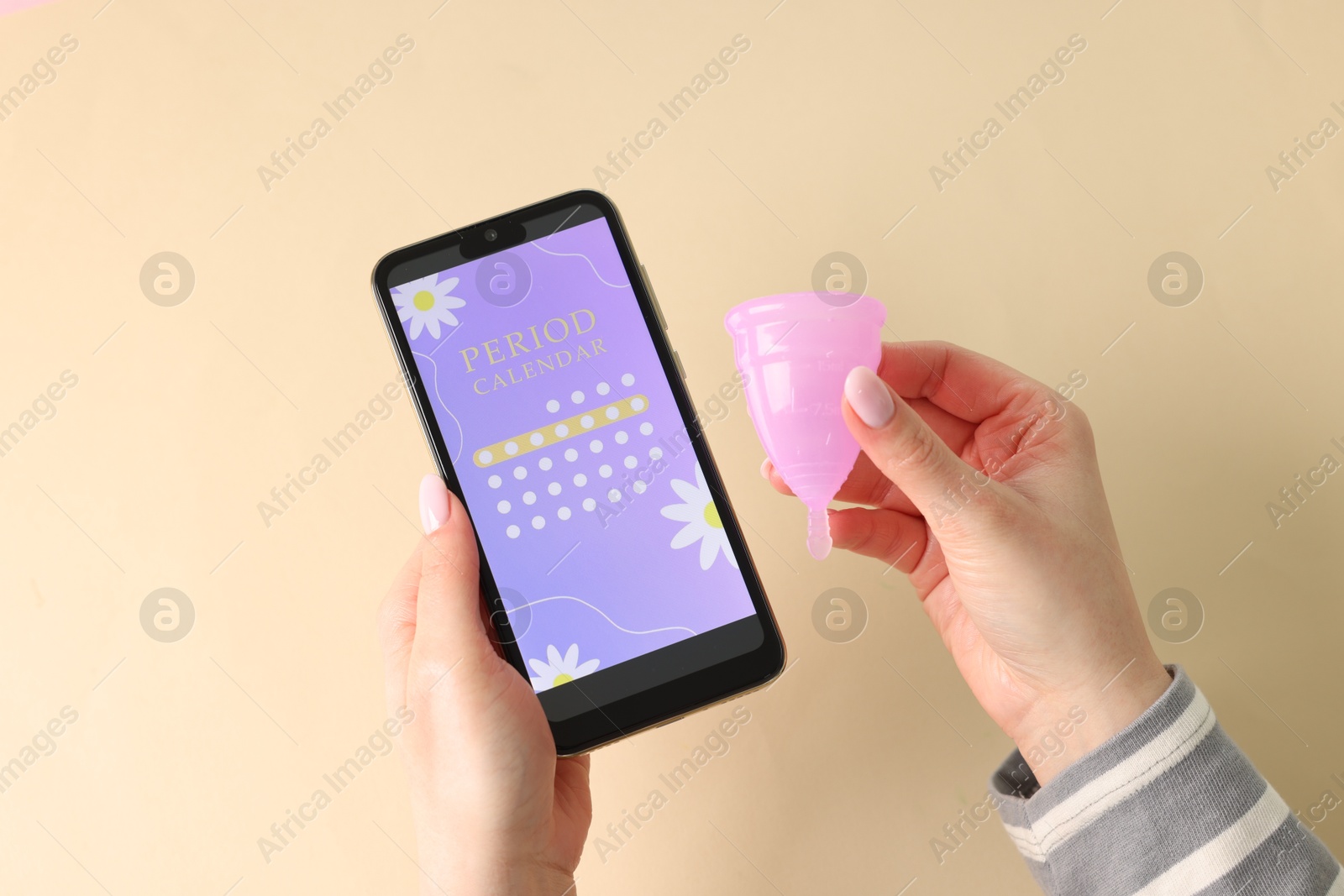 Photo of Menstruation. Woman holding smartphone with period calendar application and menstrual cup on beige background, top view