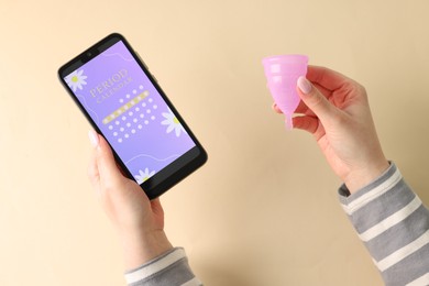 Photo of Menstruation. Woman holding smartphone with period calendar application and menstrual cup on beige background, top view