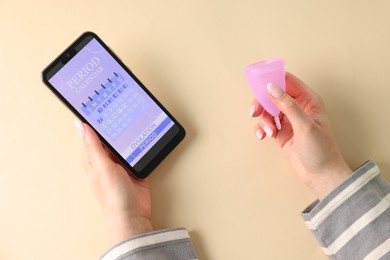 Photo of Menstruation. Woman holding smartphone with period calendar application and menstrual cup on beige background, top view