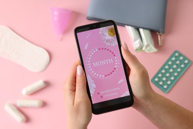 Photo of Menstruation. Woman holding smartphone with period calendar application on pink background, top view