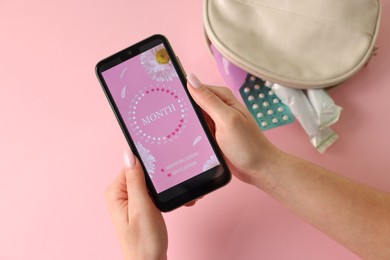 Photo of Menstruation. Woman holding smartphone with period calendar application on pink background, top view