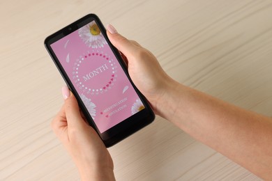 Photo of Menstruation. Woman using period calendar application on smartphone at wooden table, above view