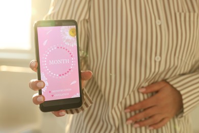 Photo of Menstruation. Woman holding smartphone with period calendar application and suffering from abdominal pain indoors, closeup