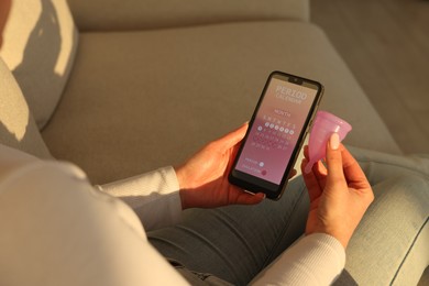 Photo of Menstruation. Woman holding smartphone with period calendar application and menstrual cup on sofa indoors, closeup