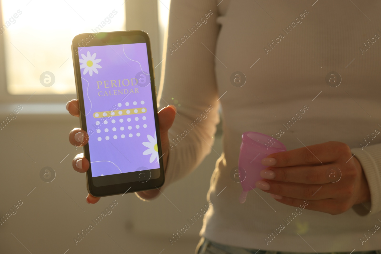 Photo of Menstruation. Woman holding smartphone with period calendar application and menstrual cup indoors, closeup