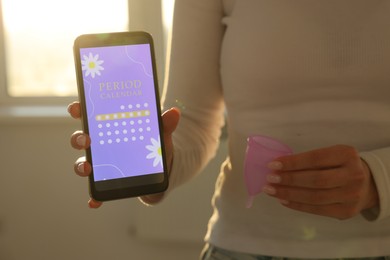 Photo of Menstruation. Woman holding smartphone with period calendar application and menstrual cup indoors, closeup