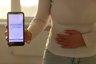 Photo of Menstruation. Woman holding smartphone with period calendar application and suffering from abdominal pain indoors, closeup