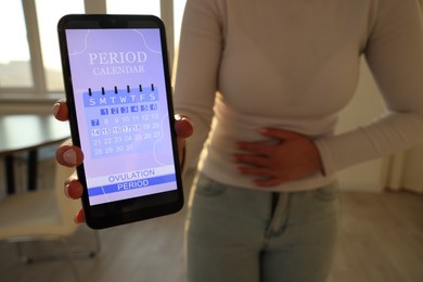 Photo of Menstruation. Woman holding smartphone with period calendar application and suffering from abdominal pain indoors, closeup