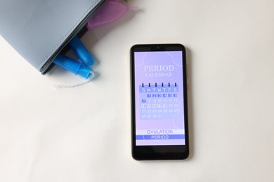 Photo of Smartphone with period calendar application and menstrual products on white table, flat lay