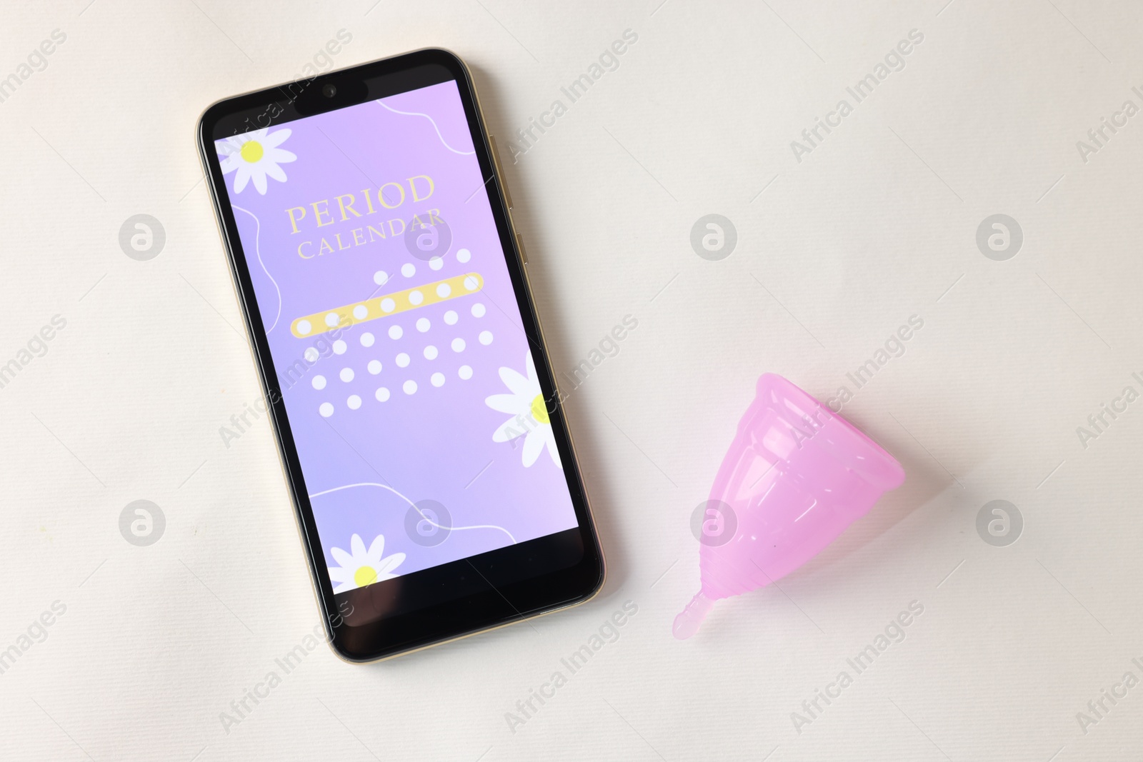 Photo of Smartphone with period calendar application and menstrual cup on white table, flat lay