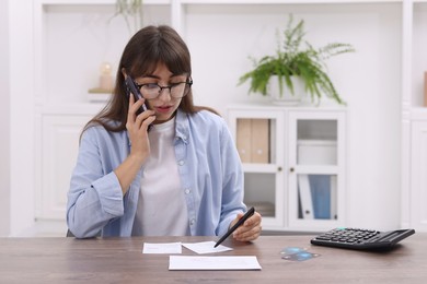 Photo of Paying bills. Woman with different invoices and calculator talking on phone at wooden table indoors