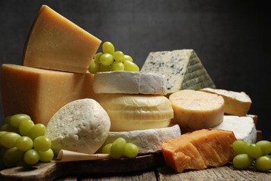 Photo of Different types of cheese and grapes on wooden table against dark background, closeup